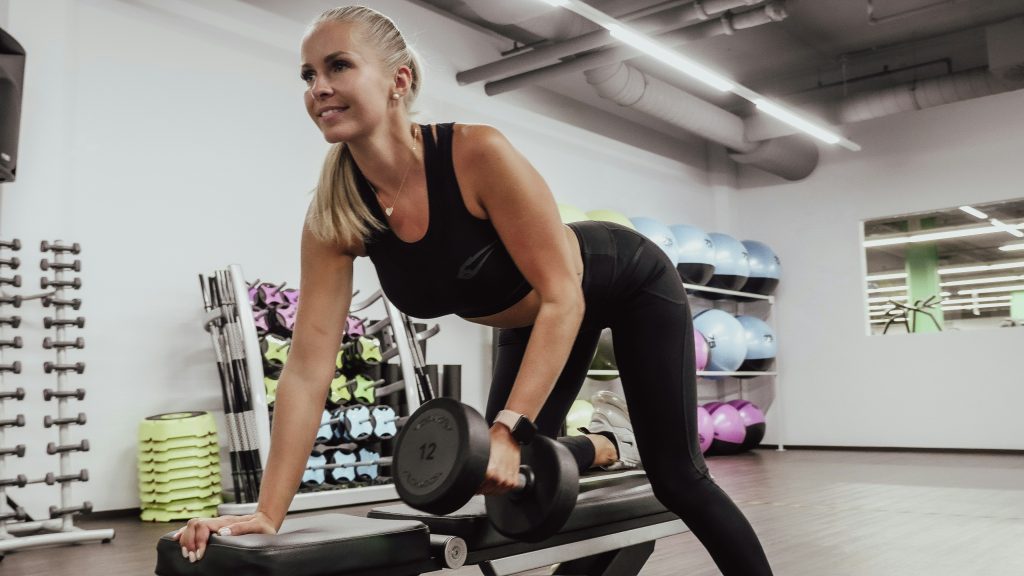 Blonde fit woman with a ponytail doing a dumbbell row on a bench in a gym.